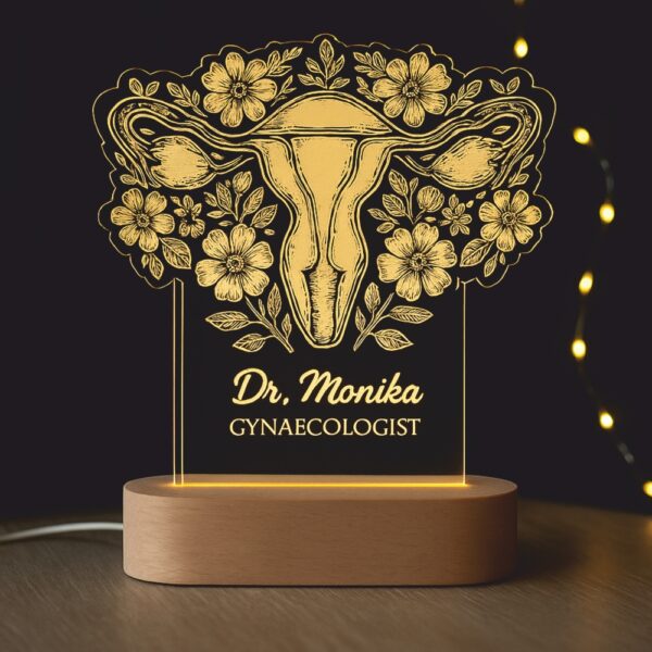 Customized LED night lamp with a floral anatomical uterus illustration and the engraved name “Dr. Monika – Gynaecologist,” glowing warmly on a wooden base in a clinic setting.