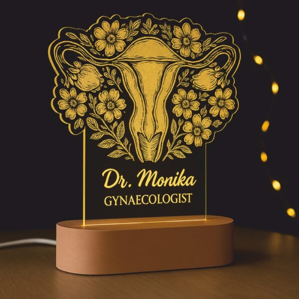 Customized LED night lamp with a floral anatomical uterus illustration and the engraved name “Dr. Monika – Gynaecologist,” glowing warmly on a wooden base in a clinic setting.