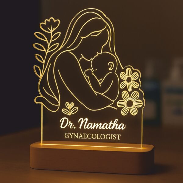 Personalized LED night lamp featuring a glowing illustration of a mother lovingly holding her baby, engraved with “Dr. Namratha – Gynaecologist,” displayed on a wooden base in a hospital setting.