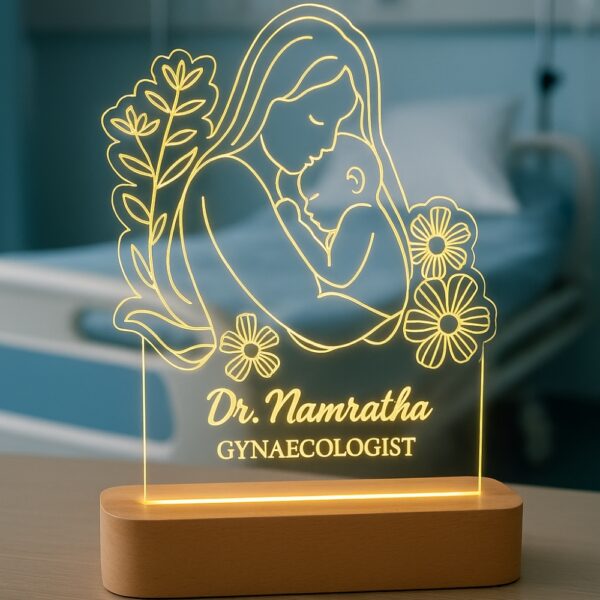 Personalized LED night lamp featuring a glowing illustration of a mother lovingly holding her baby, engraved with “Dr. Namratha – Gynaecologist,” displayed on a wooden base in a hospital setting.