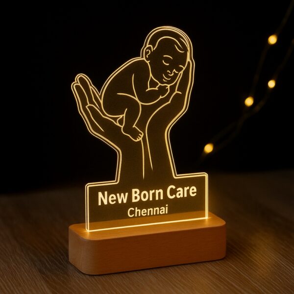 Custom LED night lamp featuring an engraved outline of a baby cradled in two hands, with the text “New Born Care – Chennai,” glowing warmly on a wooden base in a neonatal setting.