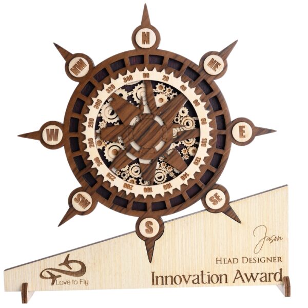 Wooden compass-themed award with intricate gear detailing, engraved base text, and nautical wheel design placed on a green pedestal.