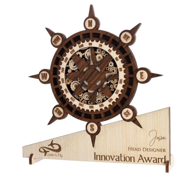 Wooden compass-themed award with intricate gear detailing, engraved base text, and nautical wheel design placed on a green pedestal.