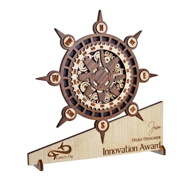 Wooden compass-themed award with intricate gear detailing, engraved base text, and nautical wheel design placed on a green pedestal.